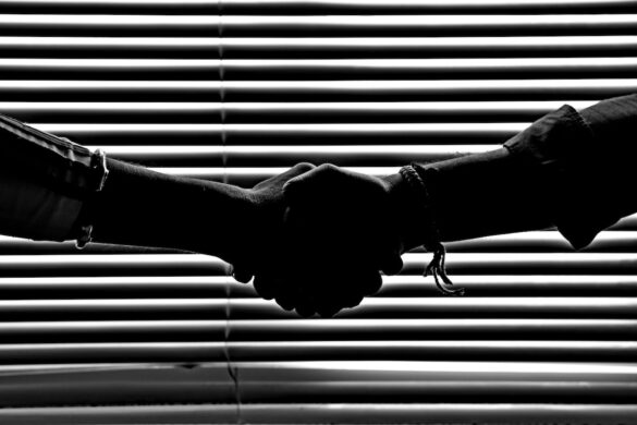 A black and white image of two hands shaking in front of a set of backlight, horizontal blinds.
