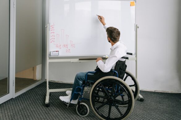A man seated in a wheelchair writes on a whiteboard in a meeting room setting.
