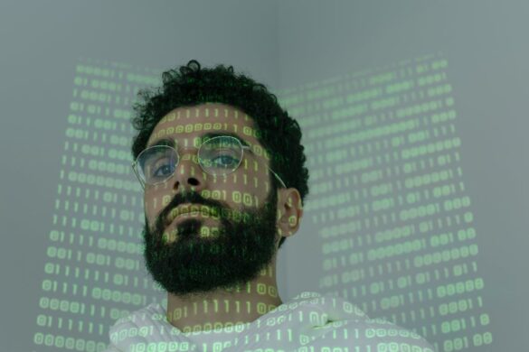 A bearded man wearing glasses looks down at a low angle camera as binary code is projected over his face and the wall behind him.