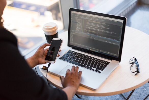 An over the shoulder shot of a woman working on a laptop which is sitting on a small table. She holds her phone in her left hand.