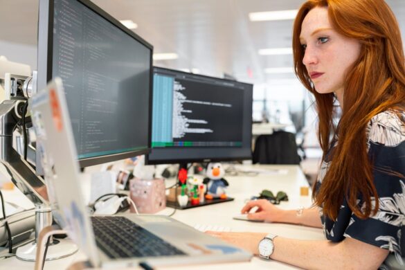 A red haired woman sits at a desk working on a laptop attached to two larger screens filled with computer code.